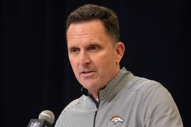 George Paton stands behind a microphone, with a black backdrop. He has on a Broncos polo.