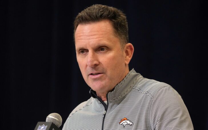 George Paton stands behind a microphone, with a black backdrop. He has on a Broncos polo.